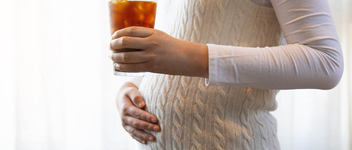 Eine Frau hält in der linken Hand ein Glas mit hellbrauner Flüssigkeit und Eiswürfeln. Die rechte Hand ruht auf ihrem schwangeren Bauch.