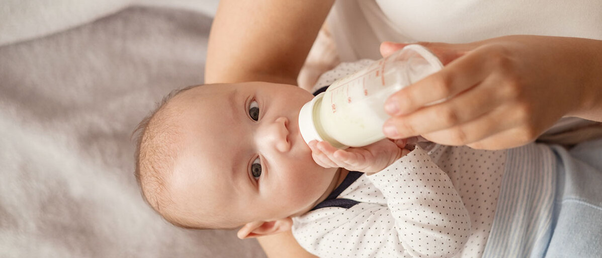 Frau hält Säugling auf dem Arm und gibt ihm eine Flasche mit Babymilch.