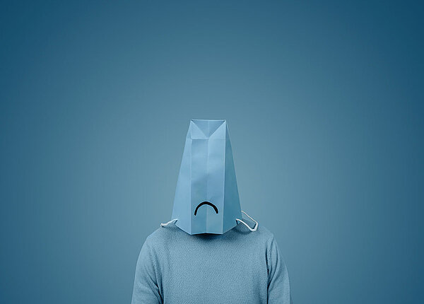 Mann mit blauer Papiertüte über dem Kopf, auf der ein trauriger Mund abgebildet ist.