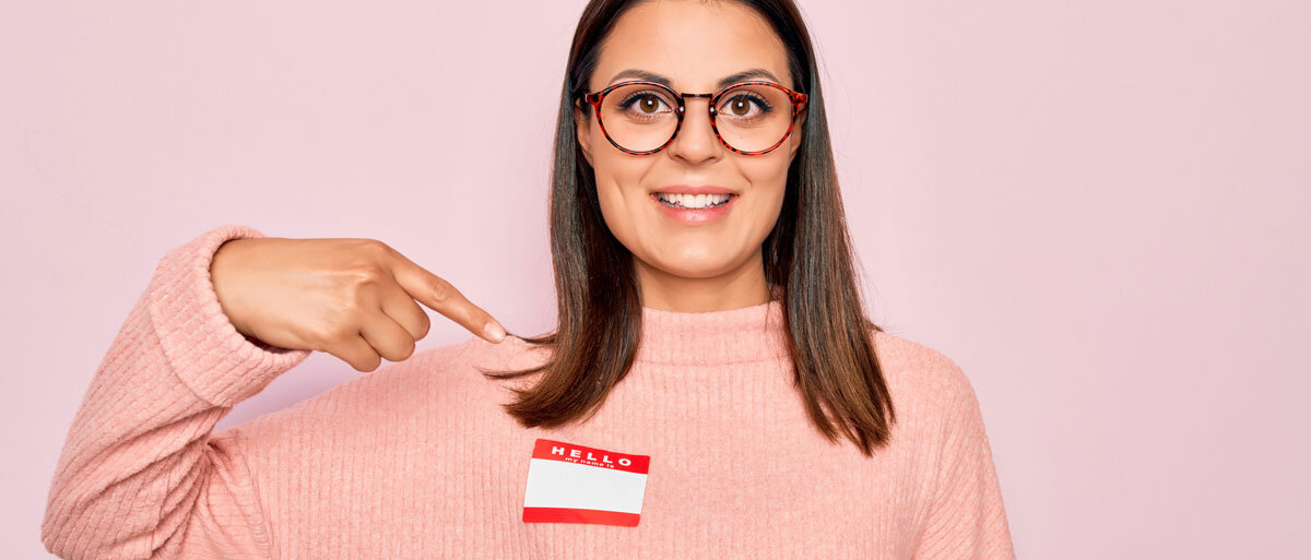 Eine Frau zeigt auf das Namensschild auf ihrem Pullover. Darauf steht "Hello my name is", aber der Name ist nicht ausgefüllt.