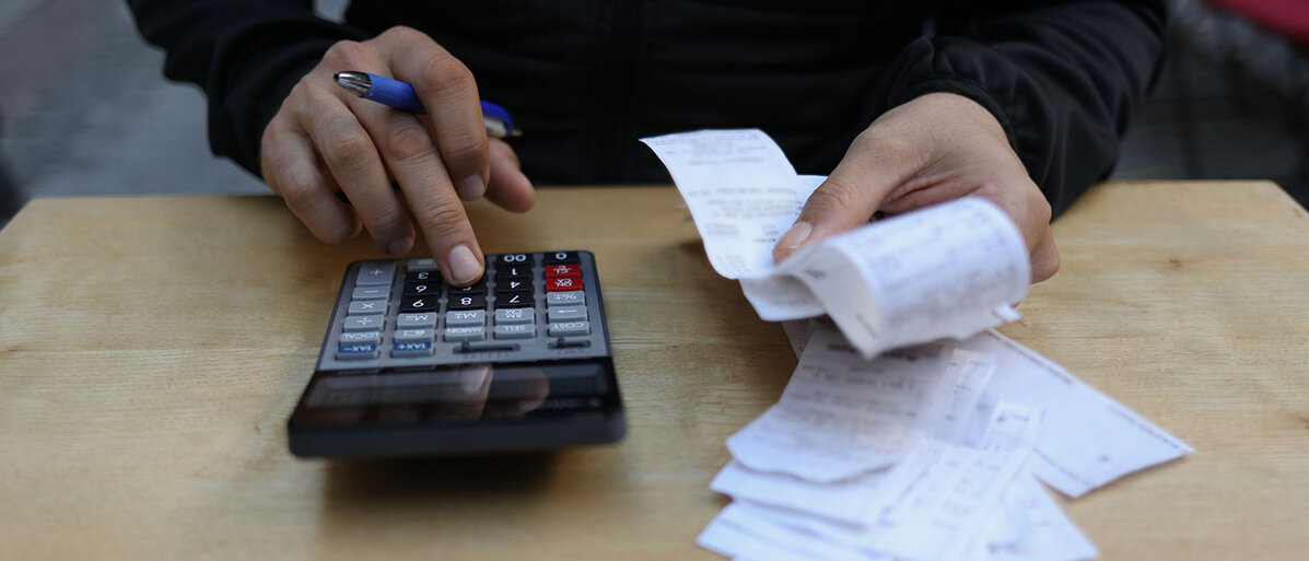Taschenrechner liegt auf einem Tisch und nebendran Rechnungsbeläge. Männer Hand gibt Beträge in Taschenrechner ein