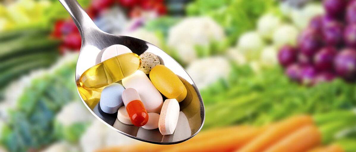 Verschiedene Tabletten und Kapseln auf einem Löffel, im Hintergrund ist Gemüse.
