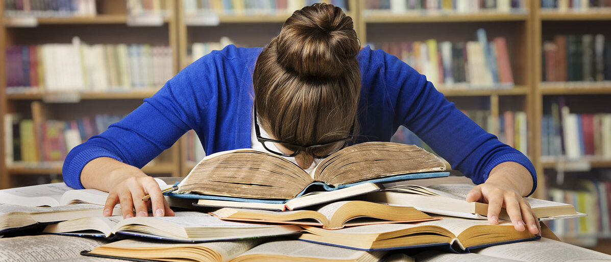 Eine Frau sitzt an einem Schreibtisch vor einem Bücherregal. Sie hat resigniert den Kopf auf einem Stapel offener Bücher abgelegt.