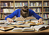 Eine Frau sitzt an einem Schreibtisch vor einem Bücherregal. Sie hat resigniert den Kopf auf einem Stapel offener Bücher abgelegt.
