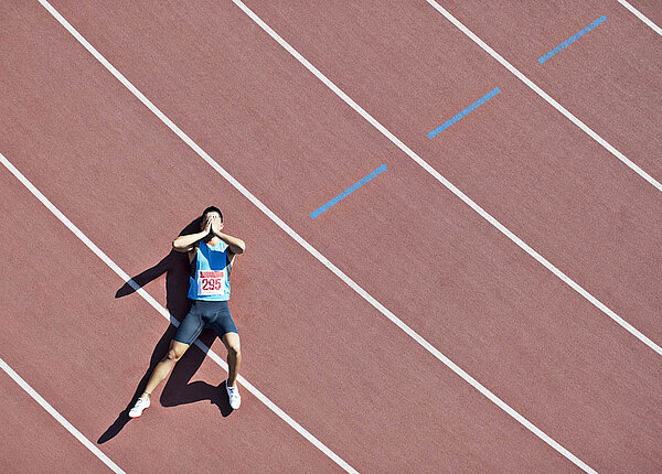 Sportliche Leistung: Wie weit kann Sport die Leistung steigern? Ein Läufer liegt erschöpft auf einer Rennbahn und schlägt die Hände vor dem Gesicht zusammen.