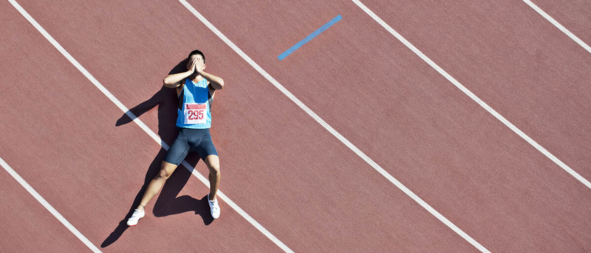 Sportliche Leistung: Wie weit kann Sport die Leistung steigern? Ein Läufer liegt erschöpft auf einer Rennbahn und schlägt die Hände vor dem Gesicht zusammen.