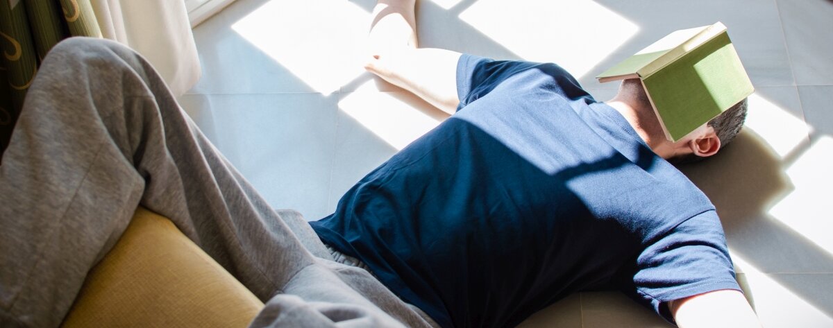 Ein Mann liegt auf dem Boden, die Füße auf dem Sofa, ein Buch über dem Gesicht.