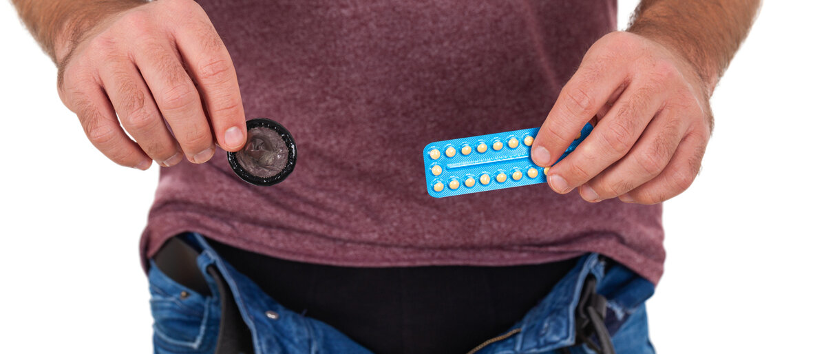 Ein Mann mit geöffnetem Gürtel hält in der einen Hand ein Kondom, in der anderen einen Pillen-Blister.