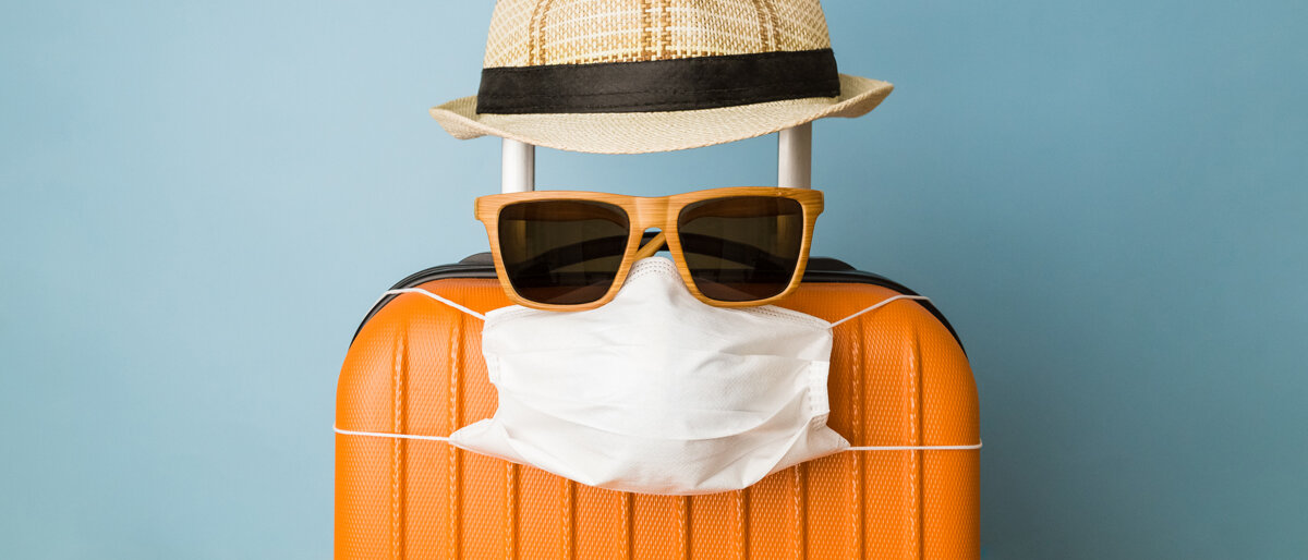 Ein Koffer trägt Sonnenhut, Sonnenbrille und Mundschutz.