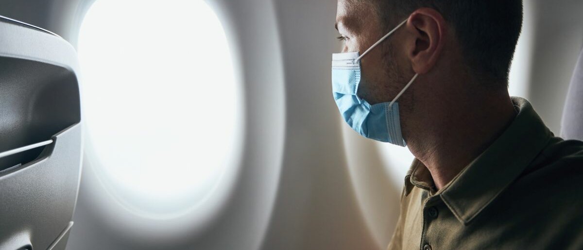 Ein Mann sitzt im Flugzeug, trägt eine Atemschutzmaske und schaut aus dem Fenster.