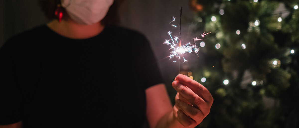 Eine Frau hält eine brennende Wunderkerze. Sie trägt Mundschutz. Im Hintergrund steht ein Weihnachtsbaum.