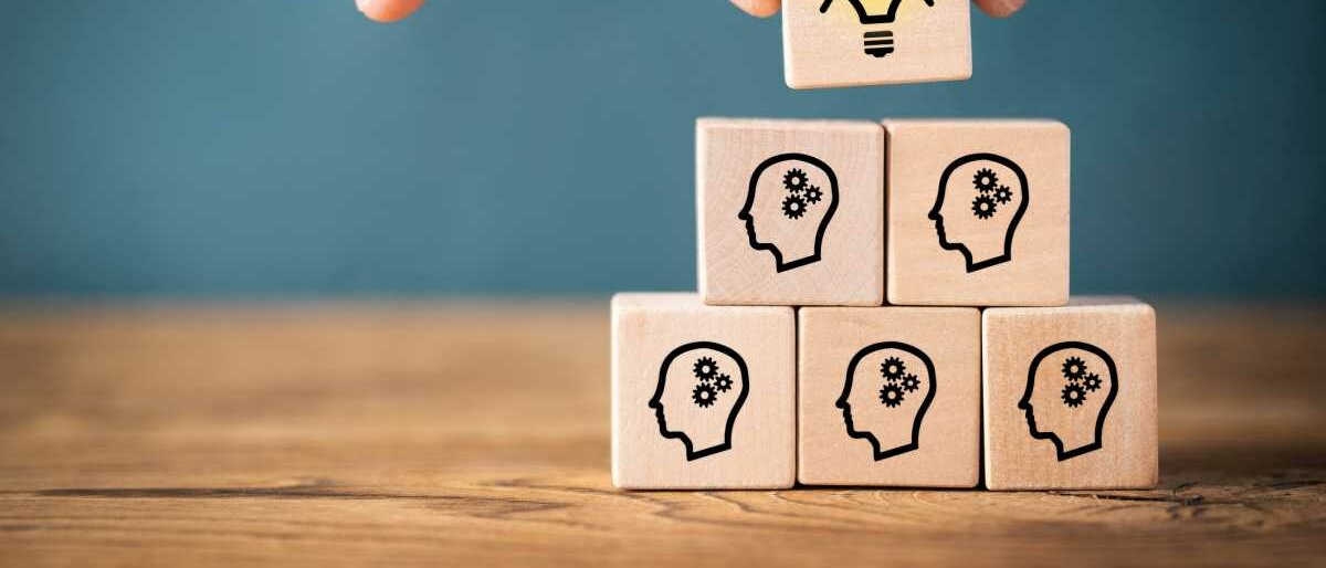 Mehrere Holzwürfel zeigen einen Kopf mit Zahnrad als Gehirn, sie stapeln sich zu einer Pyramide. Der oberste Würfel zeigt eine leuchtende Glühbirne.