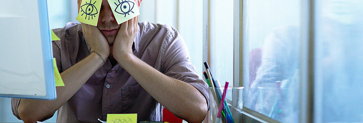 Ein Mann schläft vor seinem PC. Auf den Augen hat er Post-its mit aufgemalten offenen Augen geklebt.