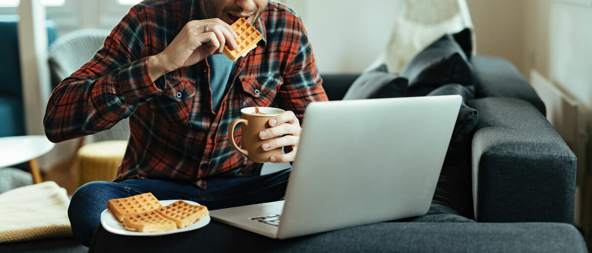 Homeoffice Ein Mann sitzt auf der Couch, schaut in seinen Laptop, isst Waffeln und trinkt aus einer Tasse.