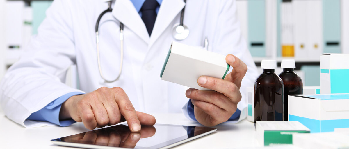 Ein Mann im Kittel hält eine Arzneimittelschachtel in der einen Hand und arbeitet mit der anderen auf einem Tablet.