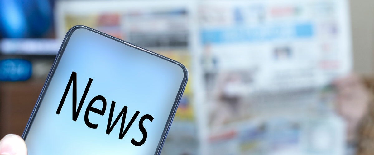 Nachrichten Im Hintergrund läuft der Fernseher, jemand hat eine Zeitung aufgeschlagen. Im Vordergrund sieht man ein Smartphone, auf dem Bildschirm steht "NEWS".