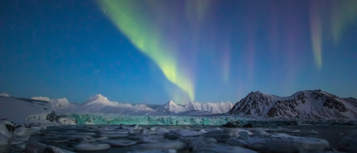 Arktis In der Dämmerung treiben lose Eisschollen vor einem Eisberg, am Himmel tanzen Polarlichter.