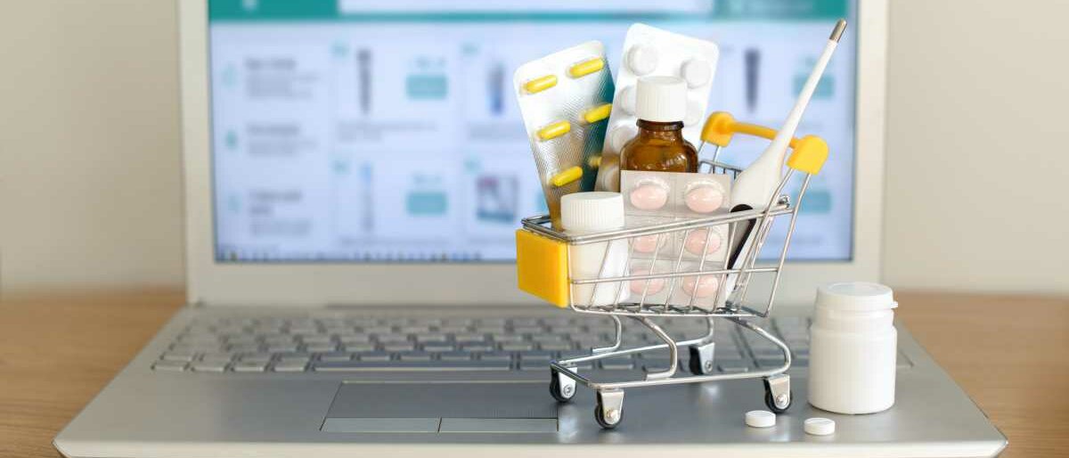 Auf der Tastatur eines aufgeklappten Laptops steht ein Spielzeug-Einkaufswagen, gefült mit Arzneimitteln. Der Bildschirm des Laptops deutet eine Online-Apotheke an.