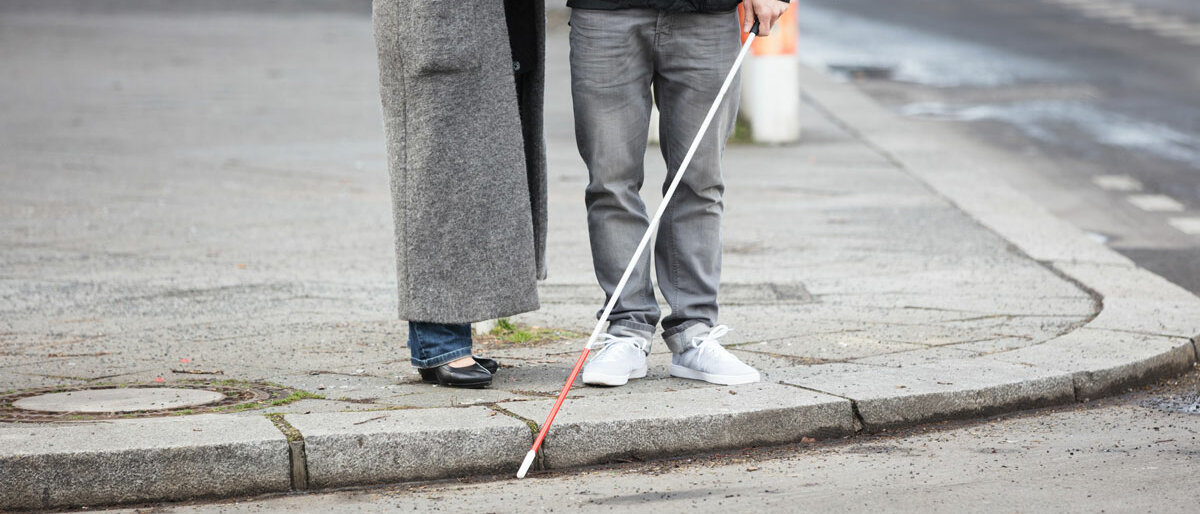 Ein sehbehinderter Mensch mit Blindenstock wird von einem anderen Menschen über eine Straße begleitet