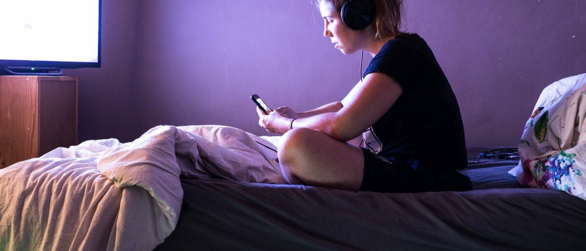 Eine junge Frau sitzt auf dem Bett, hat Kopfhörer aufgesetzt und schaut auf ihr Handy. Gleichzeitig läuft der Fernseher.