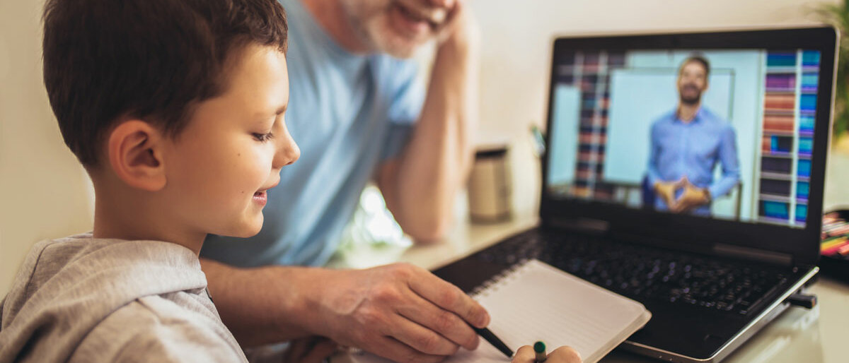 Homeschooling Ein Junge sitzt vor dem Laptop, sein Lehrer erklärt etwas in einer Videokonferenz. Der Junge macht sich Notizen, sein Vater sitzt daneben.