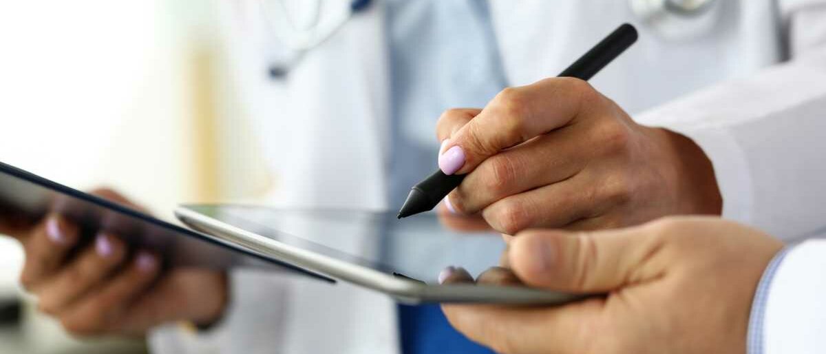 Eine Ärztin hält in der einen Hand ein Tablet, mit der anderen unterzeichnet sie digital auf einem zweiten Tablet.