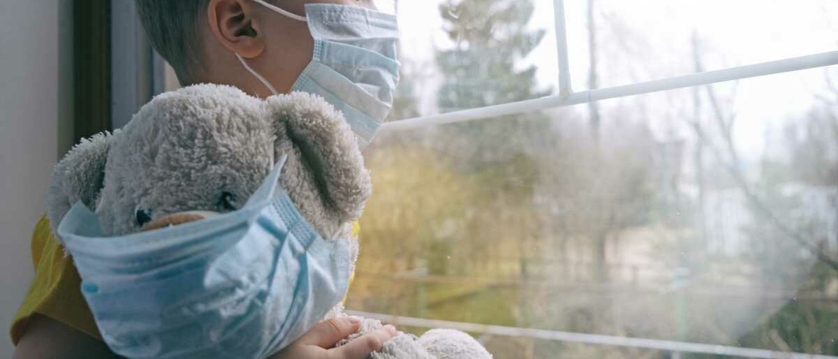Ein kleiner Junge und sein Teddy schauen aus dem Fenster, beide tragen einen Mund-Nasen-Schutz.