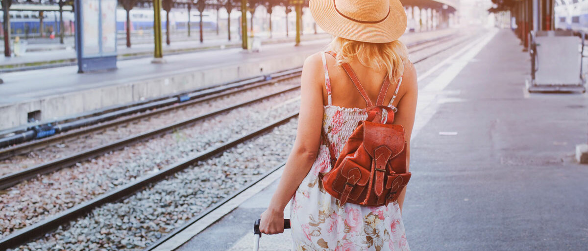 Eine Frau mit Sonnenhut, Koffer und Rucksack steht am Bahnsteig