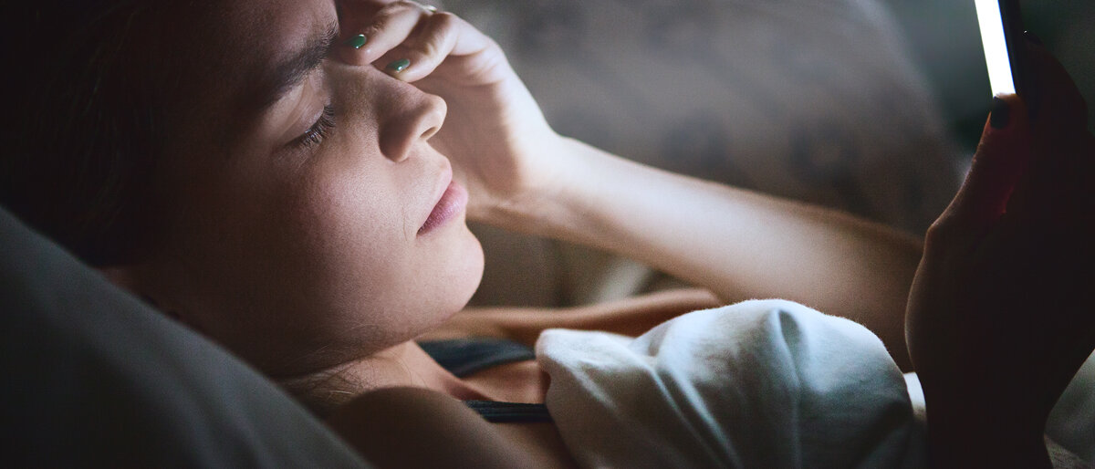 Eine Frau liegt in einem abgedunkelten Raum im Bett, hält sich den Kopf und schaut auf ihr Smartphone.