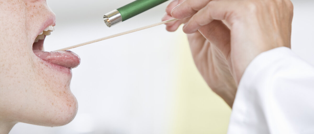 Ein Arzt oder Forscher schaut mit Hilfe eines Spatels und einer Lampe in den Rachen einer zweiten Person.