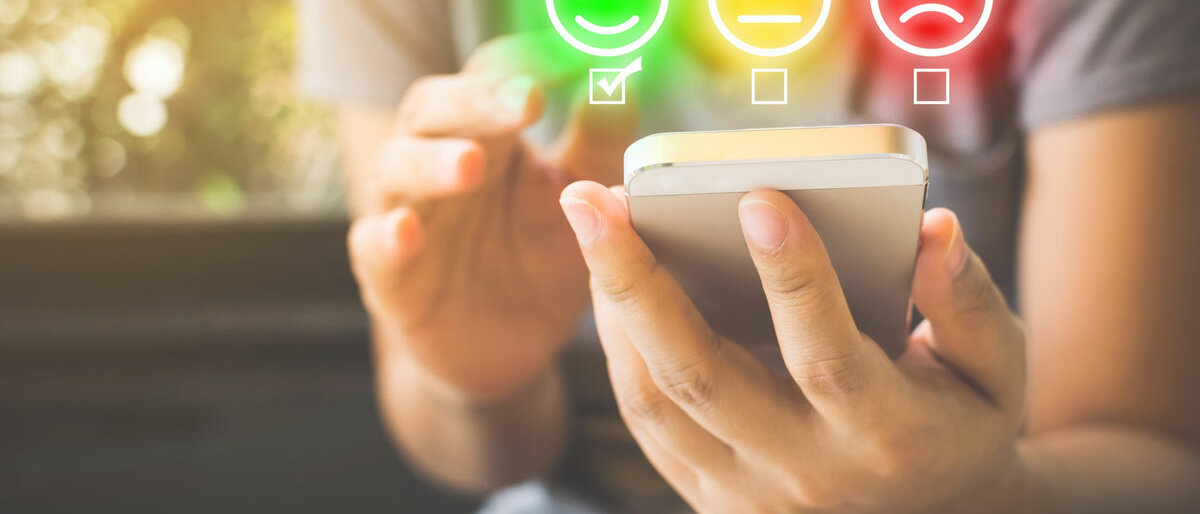 Eine Person hält ein Smartphone in der Hand. Eingeblendet sind drei Emoticons, mit denen man über zufrieden, neutral oder unzufrieden abstimmen kann.
