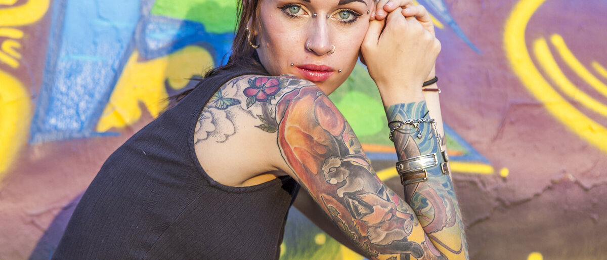 Eine junge Frau vor einer bunten Wand hat farbige Tattoos auf Arm und Rücken.
