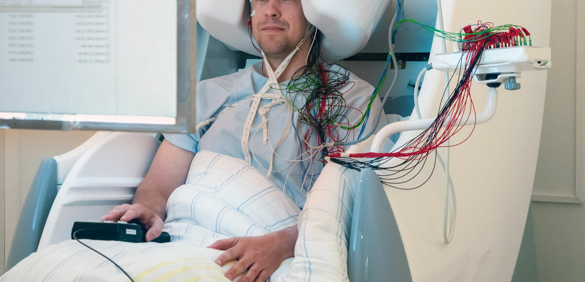 Ein Proband ist mit vielen Kabeln am Gehirn angeschlossen und sitzt in einem großen Apparat