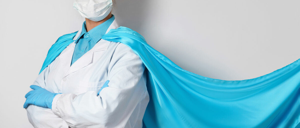 Superheld Eine Frau trägt Kittel und Mund-Nasen-Schutz, außerdem einen Superheldenumhang. Sie hat selbstbewusst die Arme veschränkt.