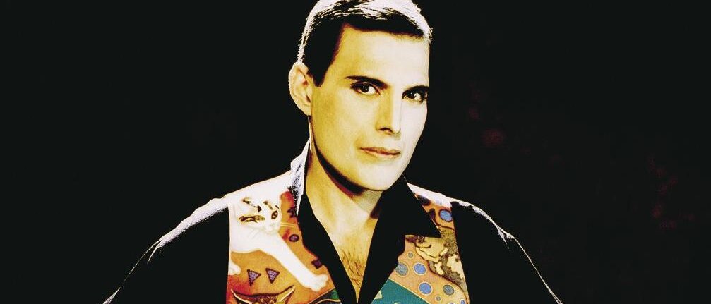 Freddie Mercury trägt eine bunte Weste mit Katzenmotiven und posiert mit selbstbewusstem Blick. 