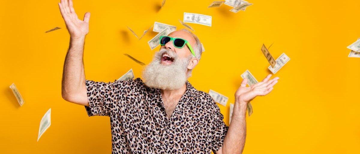 Ein älterer Mann mit Bart schmeißt Geldscheine in die Luft und freut sich.