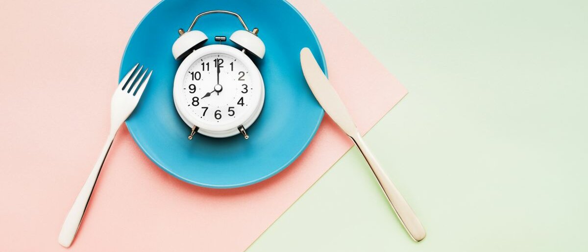 Fasten Ein blauer Teller, auf dem ein Wecker liegt, steht auf einem rosafarbenen Untergrund.