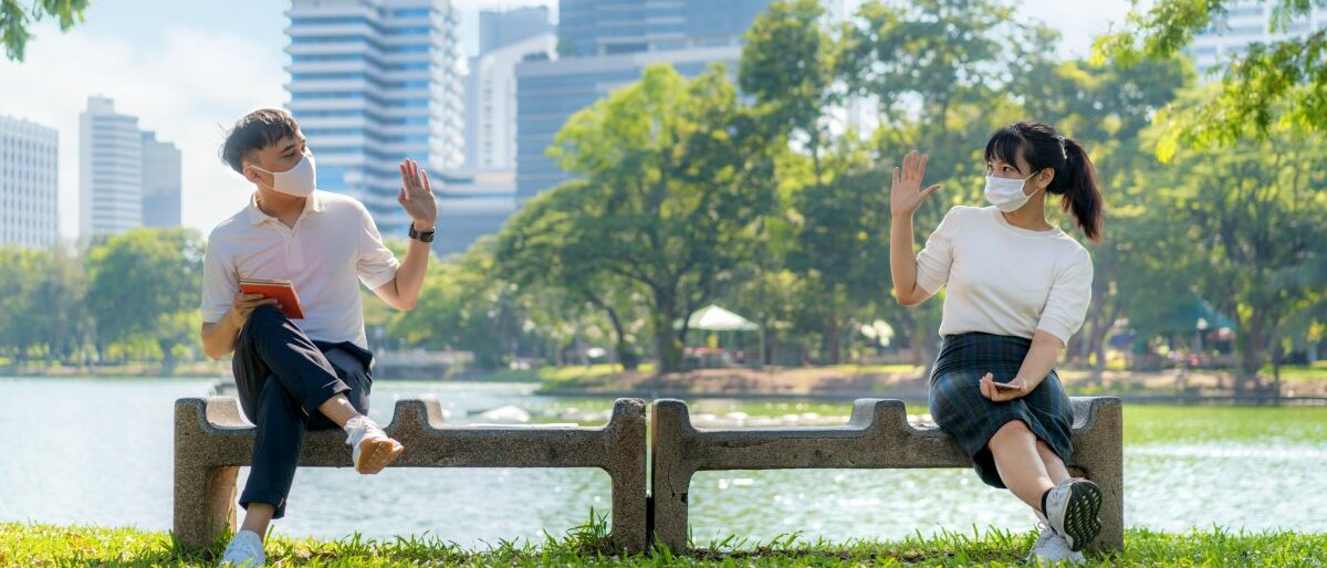 Zwei Menschen, die eine Maske tragen, auf einer Mauer sitzen und sich mit dem eingehaltenen Mindestabstand unterhalten.
