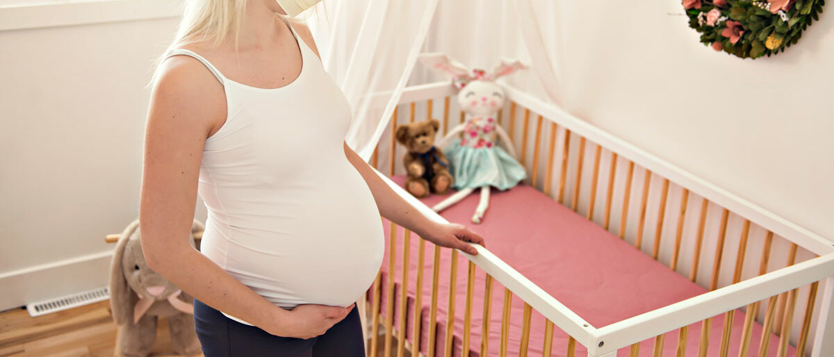eine schwangere Frau mit Mundschutz steht vor einem Kinderbett