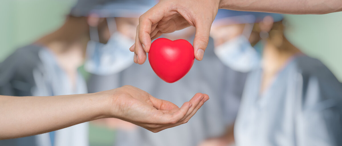 Eine Hand gibt der anderen ein Herz, im Hintergrund ein OP-Saal