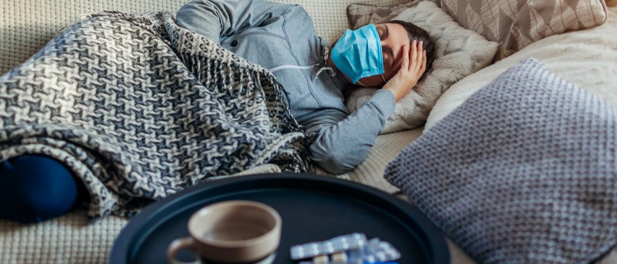 Eine Frau mit Atemschutzmaske liegt auf dem Sofa und fühlt sich schlecht.