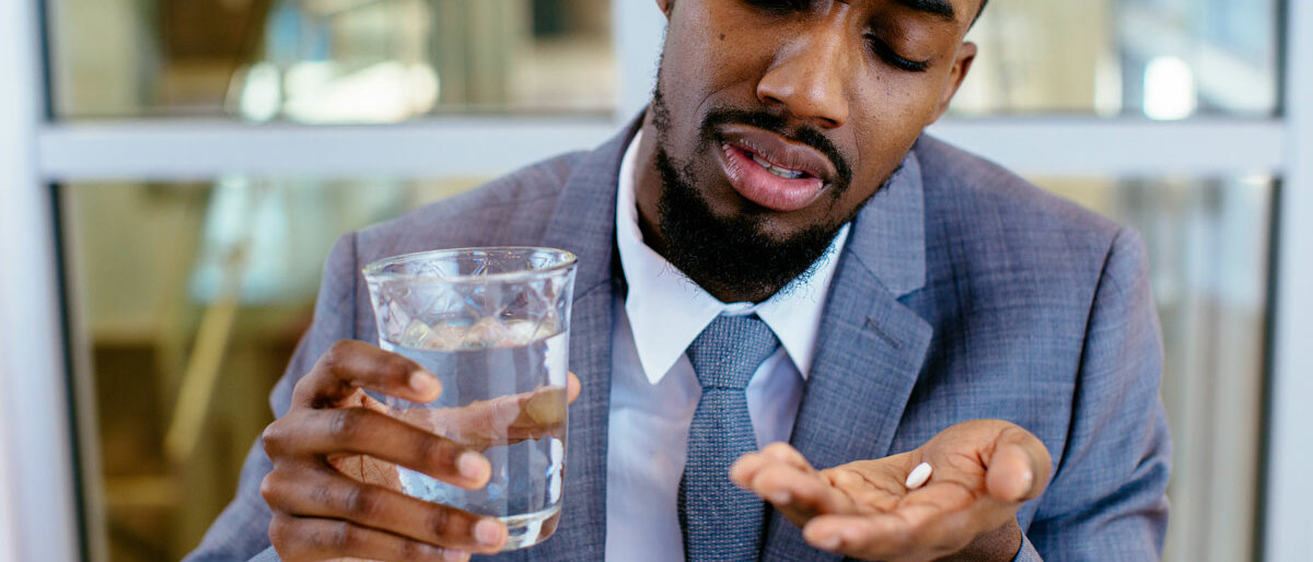 Ein Mann verzieht das Gesicht während er in der einen Hand ein Glas Wasser und in der anderen eine Tablette hält.
