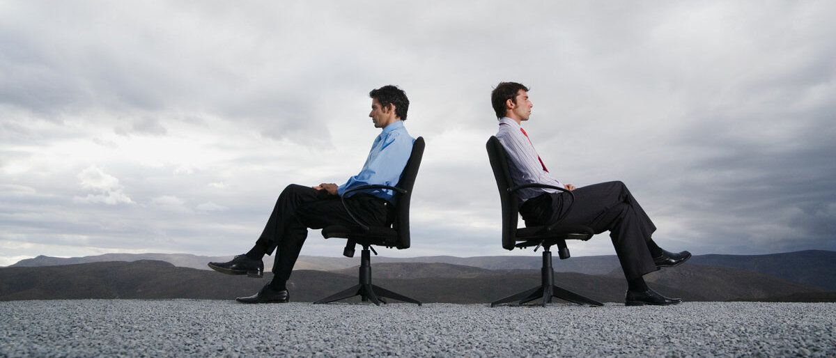 Zwei Männer sitzen in einer steinigen Landschaft auf ihren Bürostühlen. Sie wenden sich gegenseitig den Rücken zu.