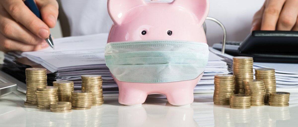 Abrechnung Ein Sparschwein mit Mundschutz steht auf einem Tisch, daneben liegen Ein-Euro-Münzen. Im Hintergrund macht ein Mann die Abrechnung.