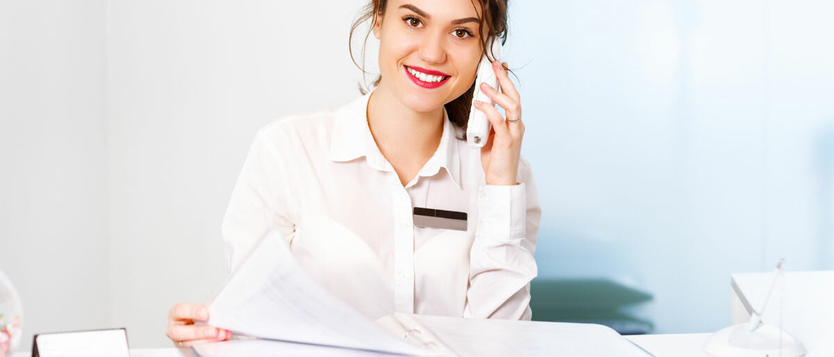Eine lächelnde Frau sitzt am Empfang und nimmt ein Telefonat entgegen