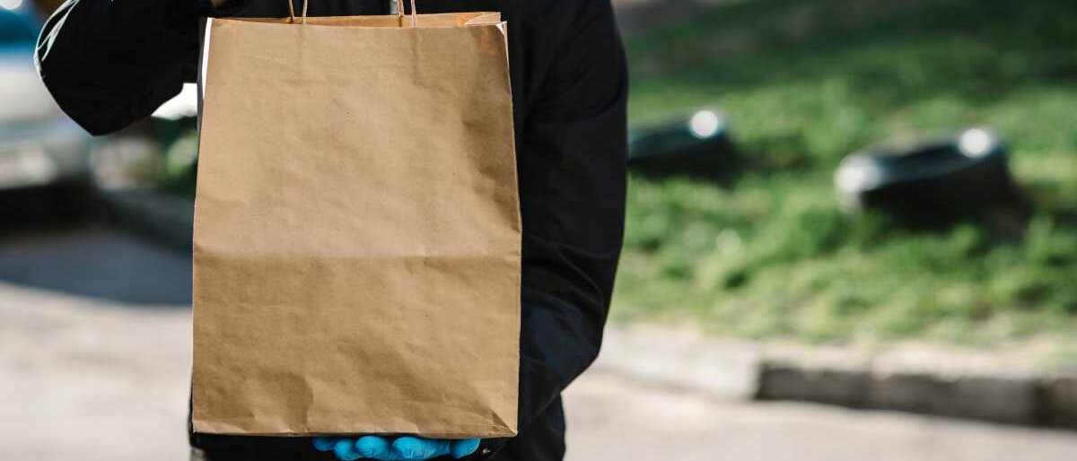 Ein Mann, der Mundschutz und Handschuhe trägt, hält eine braune Papiertüte hoch.