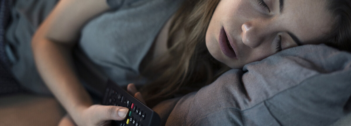 Eine junge Frau schläft auf dem Sofa mit der Fernbedienung in der Hand und wird vom Fernseher angestrahlt.