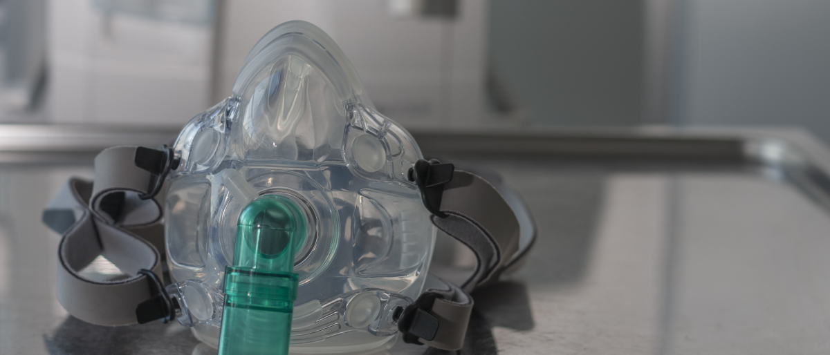 Auf einem Metalltablett liegt eine Maske zur nichtinvasiven Beatmung, im Hintergrund erkennt man ein Krankenhauszimmer.