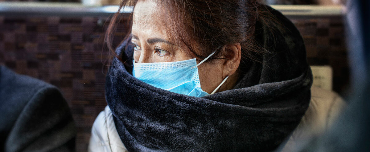 Eine Frau sitzt im Bus, eingehüllt in einen Schal und trägt einen Mundschutz