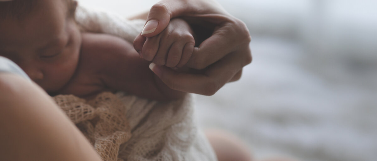 Eine Mutter hält ihr Neugeborenes im AArm, es hat ihren Finger in der Hand.